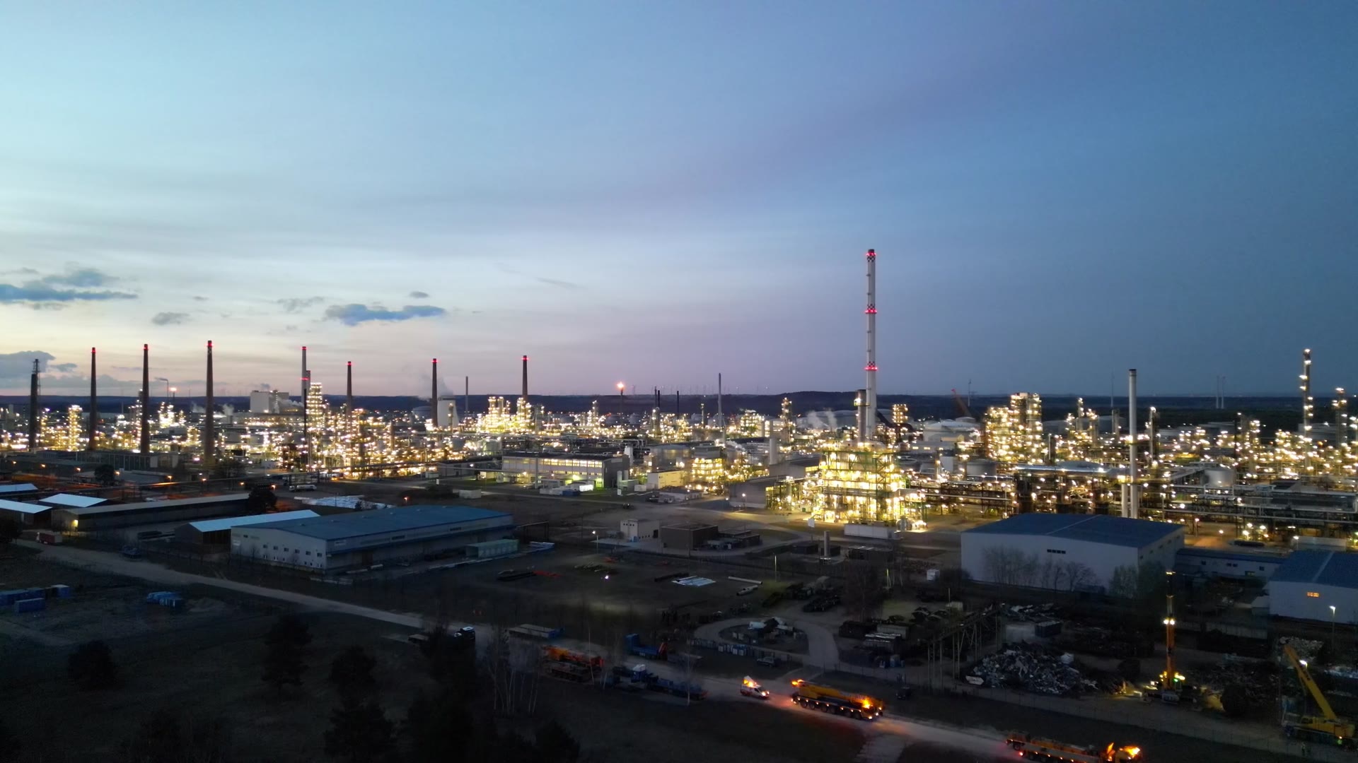 An industrial plant lit up at dusk, seen from the air, stacks and process towers against a soft sky